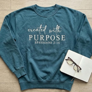 May include: Teal crewneck sweatshirt with the words "created with PURPOSE" and "EPHESIANS 2:10" in white script. A notebook and glasses rest on the right side of the sweatshirt. The sweatshirt is a solid color.