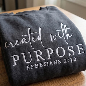 Puede incluir: Sudadera gris oscuro con texto bordado blanco que dice "created with PURPOSE" y "EPHESIANS 2:10". La sudadera doblada está sobre una superficie de madera. La sudadera es de estilo casual.