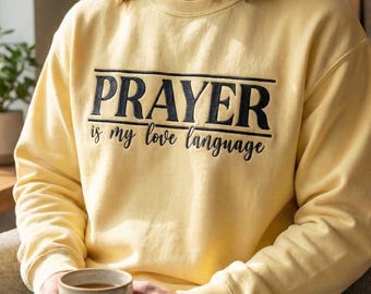 May include: A light yellow sweatshirt with the text "PRAYER is my love language" in black. The sweatshirt is long-sleeved and has a crew neck. The person is holding a beige mug.