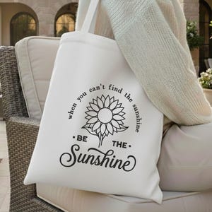 May include: A white tote bag featuring a black sunflower illustration and the words "when you can't find the sunshine, be the sunshine." The bag is held by a person wearing a cream-coloured jumper. A simple, stylish bag for carrying essentials.