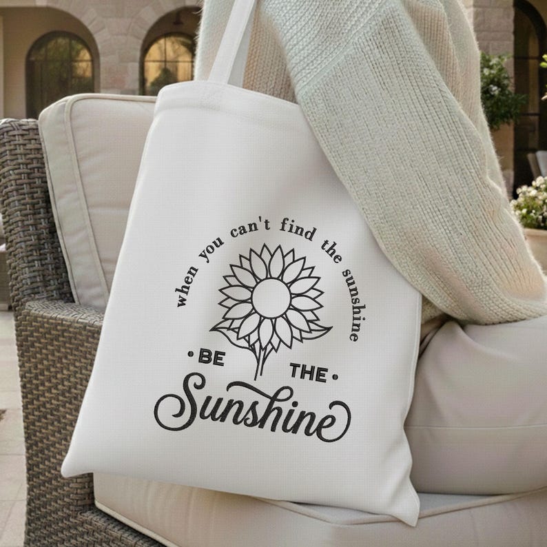 May include: A white tote bag featuring a black sunflower illustration and the words "when you can't find the sunshine, be the sunshine." The bag is held by a person wearing a cream-coloured jumper. A simple, stylish bag for carrying essentials.