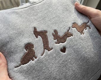 May include: A heather gray hooded sweatshirt featuring brown embroidery of four dachshunds in various poses, including one sitting, one standing on hind legs, one jumping, and one walking, accompanied by a small embroidered brown heart.