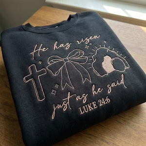May include: Black sweatshirt with embroidered design. The design includes a cross, a bow, and the text "He has risen just as he said Luke 24:6" in a light brown color. The sweatshirt is folded on a wooden surface.