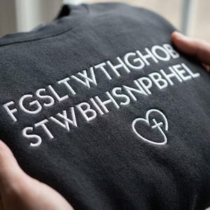 以下が含まれることがあります： FGSLTWTHGHOB STWBIHSNPBHELという白い刺繍文字と、中に小さな十字架のシンボルが入った白い刺繍のハートアイコンが胸の右下に施された、チャコールグレーのスウェットシャツ。