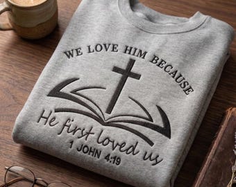 May include: A gray sweatshirt with the embroidered text "WE LOVE HIM BECAUSE He first loved us 1 JOHN 4:19" and a cross over an open book design. The sweatshirt is folded on a wooden surface.
