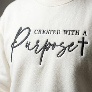 Puede incluir: Sudadera de color crema con el texto bordado "CREATED WITH A Purpose" en escritura gris oscuro y una cruz. La sudadera está hecha de un material suave.