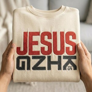 May include: A folded beige sweatshirt with the words "JESUS IS" embroidered in red and black thread. The sweatshirt is held by two hands, showcasing the design prominently.
