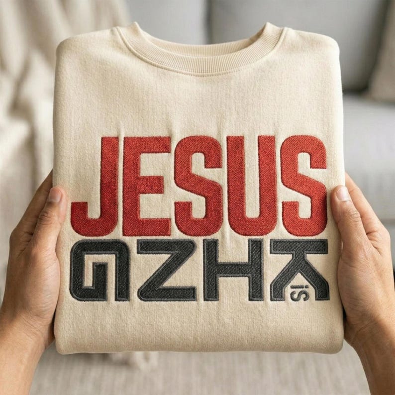 May include: A folded beige sweatshirt with the words "JESUS IS" embroidered in red and black thread. The sweatshirt is held by two hands, showcasing the design prominently.
