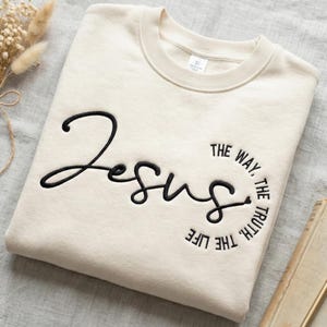 May include: Cream-colored sweatshirt with the word "Jesus" embroidered in black cursive script. The phrase "The Way, The Truth, The Life" is embroidered in a circular pattern around the word Jesus. The sweatshirt has a crew neck.