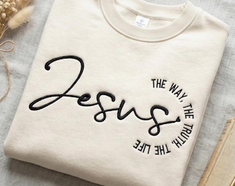May include: Cream-colored sweatshirt with the word "Jesus" embroidered in black cursive script. The phrase "The Way, The Truth, The Life" is embroidered in a circular pattern around the word Jesus. The sweatshirt has a crew neck.