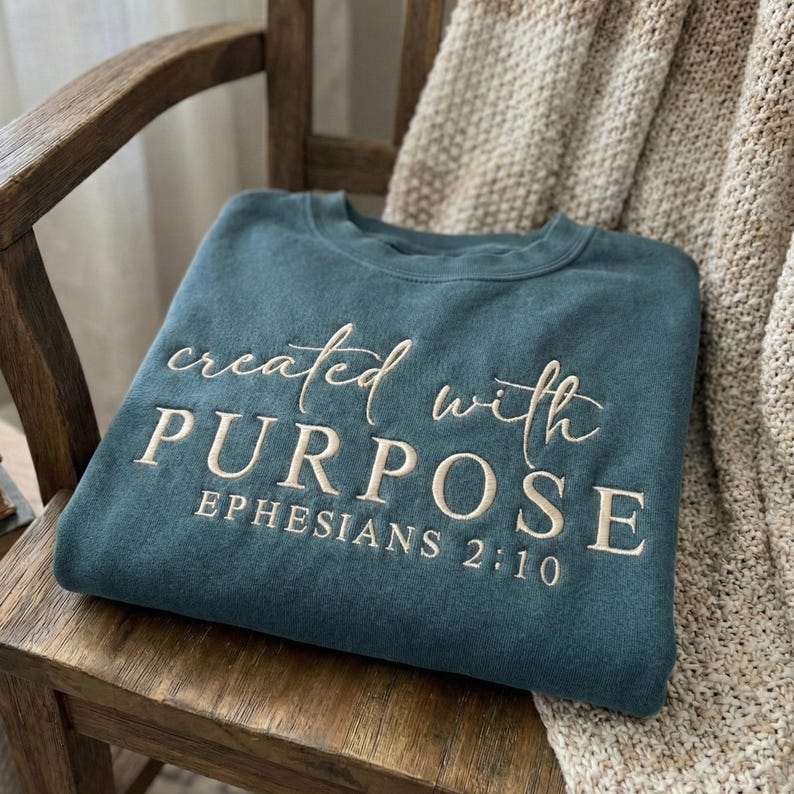 May include: Teal-colored sweatshirt with the embroidered text "created with PURPOSE" and "EPHESIANS 2:10" in a cream color. The sweatshirt is folded on a wooden chair with a neutral-toned blanket in the background.