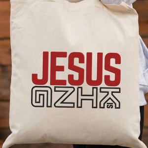 May include: Cream-colored tote bag with the word "JESUS" in bold red letters and the letters "GZHK" in a black outlined font. The bag is made of a sturdy fabric.