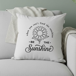 May include: White square cushion featuring a black sunflower design and the text "when you can't find the sunshine, be the sunshine". The cushion is on a grey sofa with a white blanket.