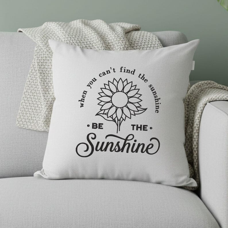 May include: White square cushion featuring a black sunflower design and the text "when you can't find the sunshine, be the sunshine". The cushion is on a grey sofa with a white blanket.