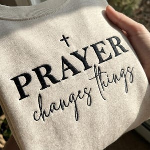 May include: A beige crewneck sweatshirt featuring black embroidered text that reads PRAYER changes things, with a small cross symbol positioned above the word PRAYER. The garment has a ribbed neckline and a soft, textured fabric finish.