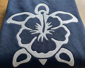 May include: A navy blue sweatshirt with a white embroidered hibiscus flower design. The flower is centered on the front of the folded sweatshirt. The sweatshirt is made of a soft material and is folded on a wooden surface.