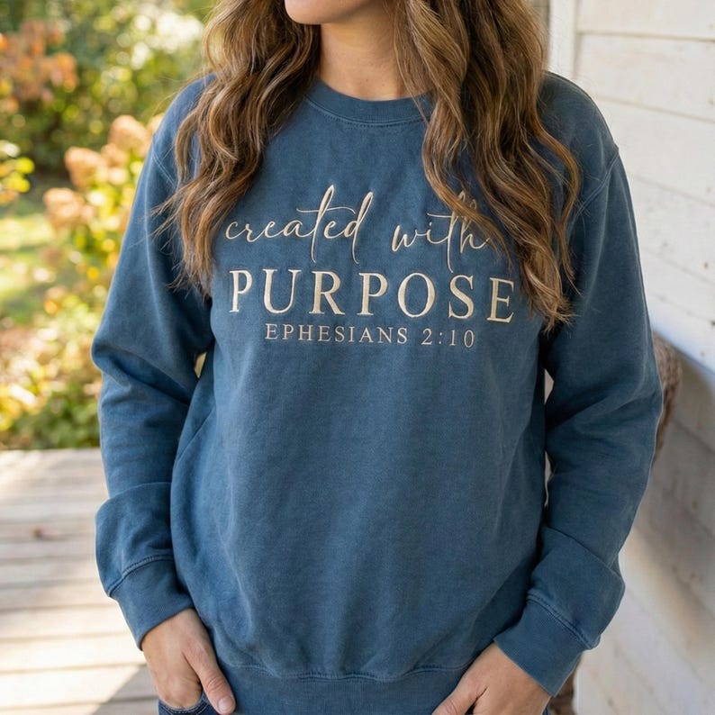 May include: A blue crew neck sweatshirt with the words "created with PURPOSE" and "EPHESIANS 2:10" printed in a light tan color. The sweatshirt has long sleeves and a relaxed fit.