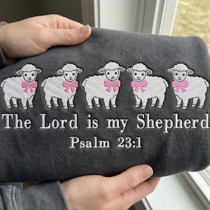 May include: Dark gray sweatshirt embroidered with five white sheep wearing pink bows. The text "The Lord is my Shepherd Psalm 23:1" is embroidered below the sheep. The sweatshirt is held by a person.