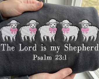 May include: Dark gray sweatshirt embroidered with five white sheep wearing pink bows. The text "The Lord is my Shepherd Psalm 23:1" is embroidered below the sheep. The sweatshirt is held by a person.