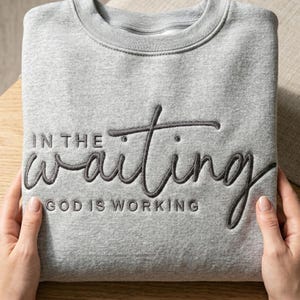 May include: A gray sweatshirt with the embroidered text "IN THE waiting GOD IS WORKING" in a dark gray script. The sweatshirt is folded and held by two hands.