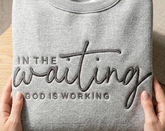 May include: A gray sweatshirt with the embroidered text "IN THE waiting GOD IS WORKING" in a dark gray script. The sweatshirt is folded and held by two hands.