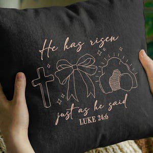 May include: Black square pillow with a light brown outline design featuring a cross, bow, and empty tomb. The text "He has risen just as he said Luke 24:6" is written in a cursive font.