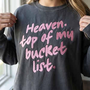 May include: Dark gray sweatshirt with the embroidered phrase "Heaven. top of my bucket list." The text is in a soft pink color. The sweatshirt has a vintage, washed appearance.