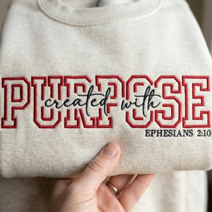 May include: A cream-coloured sweatshirt featuring the word "PURPOSE" embroidered in red block letters. The phrase "created with" is in a cursive font, and "Ephesians 2:10" is below. The sweatshirt is held by a hand.