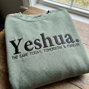 Puede incluir: Sudadera verde salvia con la palabra "Yeshua" bordada en negro, junto con la frase "THE SAME TODAY, TOMORROW & FOREVER". La sudadera está doblada sobre una superficie de madera, cerca de una ventana.