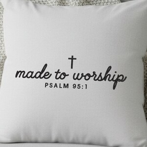 May include: A square, light gray pillow with the words "made to worship" in a black script font, a black cross, and the text "PSALM 95:1" in a smaller font. The pillow is made of a textured fabric.