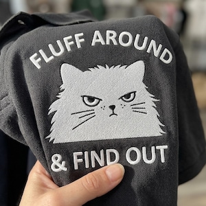 May include: Dark gray polo shirt with white embroidered text and a cat face. The text reads "FLUFF AROUND & FIND OUT". The cat has an angry expression.