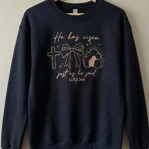 May include: Black sweatshirt with embroidered text and graphics. The design includes a cross, a bow, and an empty tomb, with the words "He has risen just as he said" and the Bible verse Luke 24:6.