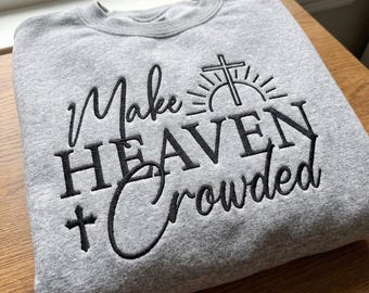 May include: A gray sweatshirt with the embroidered phrase "Make Heaven Crowded" in black script and block letters. The design includes a cross and a sunburst. The sweatshirt is folded neatly on a wooden surface.