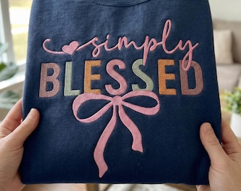 May include: Navy blue sweatshirt with the embroidered text "simply BLESSED" in pink, orange, green, and brown. A pink bow is embroidered below the text. The sweatshirt is held by two hands.