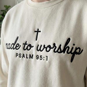 May include: Cream-colored sweatshirt with the embroidered text "made to worship" in black script, a black cross, and the text "PSALM 95:1". The sweatshirt has a crew neck.