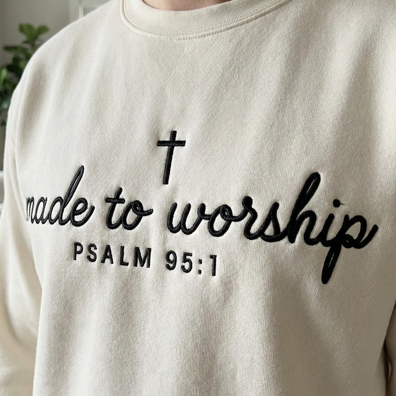 May include: Cream-colored sweatshirt with the embroidered text "made to worship" in black script, a black cross, and the text "PSALM 95:1". The sweatshirt has a crew neck.