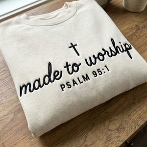 May include: Cream-colored sweatshirt with the embroidered text "made to worship" and a cross symbol. The text "PSALM 95:1" is also embroidered in black. The sweatshirt is folded on a wooden surface.