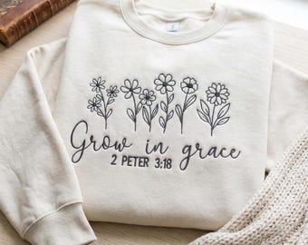 May include: Cream-colored sweatshirt with embroidered black floral design and the words "Grow in grace" with the reference "2 Peter 3:18". The sweatshirt is folded on a wooden surface with a book and a knitted blanket.