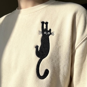 以下が含まれることがあります： クリーム色のスウェットシャツ。黒い刺繍の猫のデザインが施されています。猫は逆さまにぶら下がっており、足が伸びています。スウェットシャツはクルーネックで長袖です。