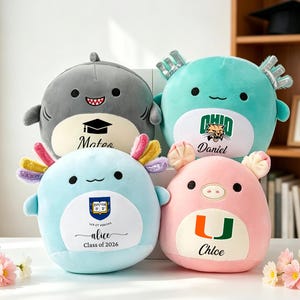 May include: Four plush toys in the shape of a shark, axolotls, and a pig. The shark is gray, the axolotls are blue and teal, and the pig is pink. Each has a name and a logo or graduation cap printed on it.