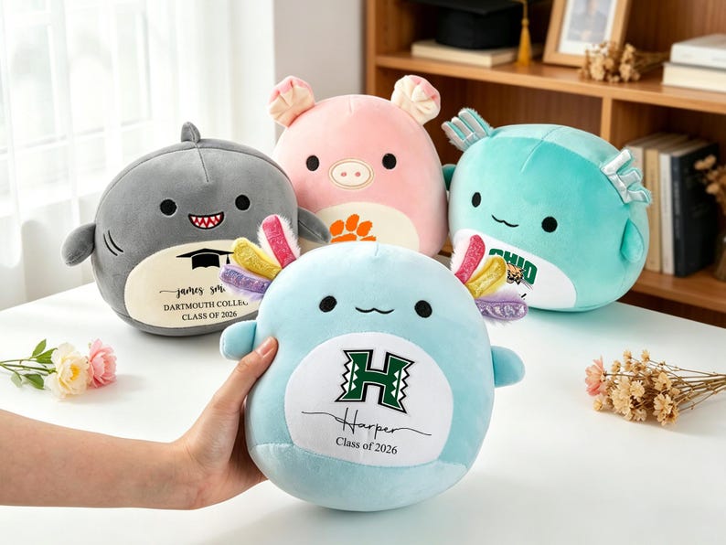 May include: Four plush toys in the shape of animals are displayed. A gray shark, a pink pig, a light blue axolotl, and a teal fish. The axolotl is held by a hand. Each toy has a college logo and name. The shark says "Dartmouth College Class of 2026".