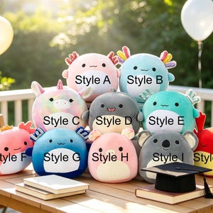 May include: A collection of plush toys in various styles and colors, including pink, blue, and gray. Each plush has a unique design, such as an axolotl, shark, and koala. The toys are arranged on a wooden table with books and a graduation cap.