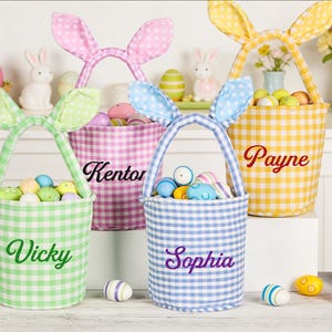 May include: Four Easter baskets in pastel shades, each with bunny ear handles and a name embroidered on the front. The baskets are filled with colourful Easter eggs. A decorative bunny and Easter eggs are also visible.