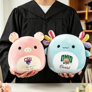 May include: Two plush toys: a pink pig and a blue axolotl. The pig has "CSU", "Harper", and "Class of 2026" printed on it. The axolotl has "OHIO" and "Daniel" printed on it. Both are held by a person wearing a graduation gown.