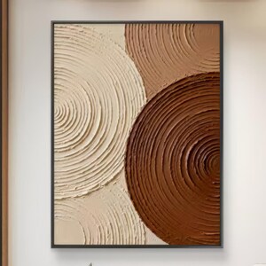 May include: Abstract wall art featuring a textured design of overlapping circles in shades of cream, tan, and brown. The artwork is framed in a black border and hangs on a white wall. The room has wooden furniture and a modern aesthetic.