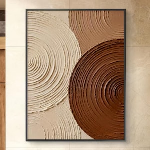 May include: Abstract wall art featuring a black framed canvas with a textured design. The artwork showcases three overlapping circular shapes in shades of beige, tan, and brown. The textured surface creates a sense of depth and visual interest.