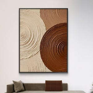 May include: Abstract wall art featuring a textured design of overlapping circles in shades of cream, tan, and brown. The artwork is framed in a black rectangular frame and hangs on a white wall.