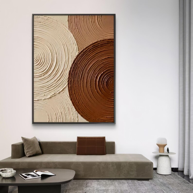 May include: Abstract wall art featuring a textured design of overlapping circles in shades of cream, tan, and brown. The artwork is framed in a black rectangular frame and hangs on a white wall.
