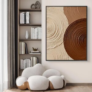 May include: Abstract wall art featuring textured concentric circles in shades of cream, tan, and brown. The artwork is framed in black and hangs on a neutral-colored wall. The room also includes a white cloud-shaped chair and a bookshelf filled with books.