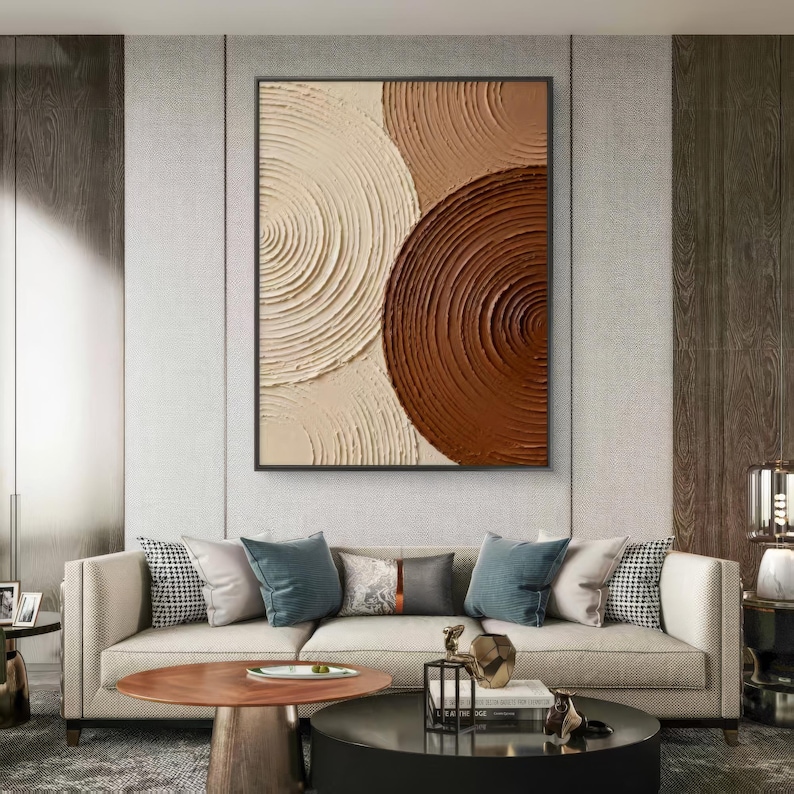 May include: Abstract wall art featuring a framed canvas with textured, concentric circles in shades of cream, tan, and brown. The artwork hangs above a beige sofa with teal and patterned throw pillows, complemented by a round coffee table.